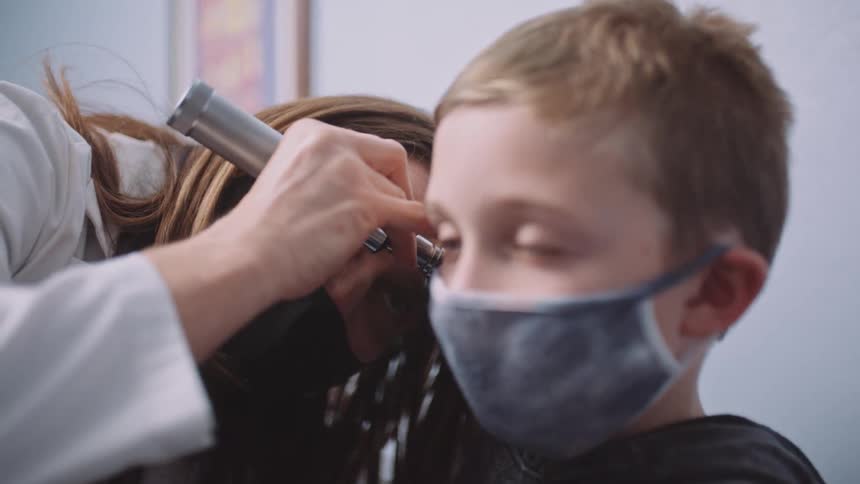 A doctor with a mask and a lab coat in a clinic looking into a boys ear
