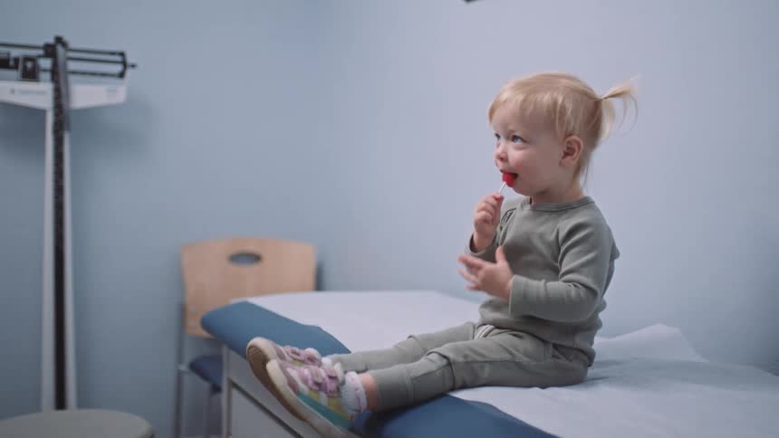 A young girl kicks her legs as she eats a lollipop while sitting on an examination table in a doctors office