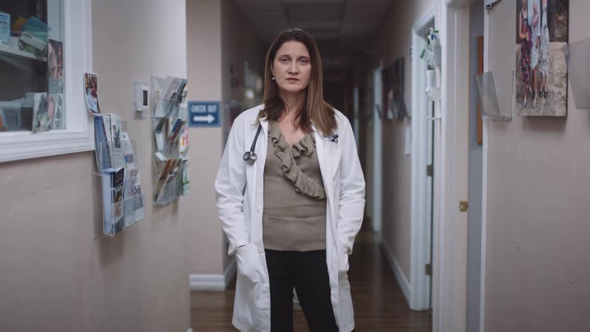 A doctor poses with her hands in her white coat while standing in the hallway at her clinic