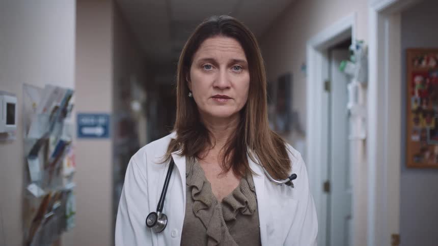 A female doctor stands in a clinic hallway with exam rooms behind her staring straight ahead