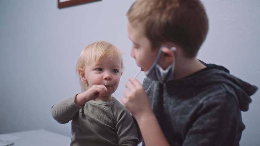 A boy holds onto his sister as they sit on a hospital bed eating candy suckers