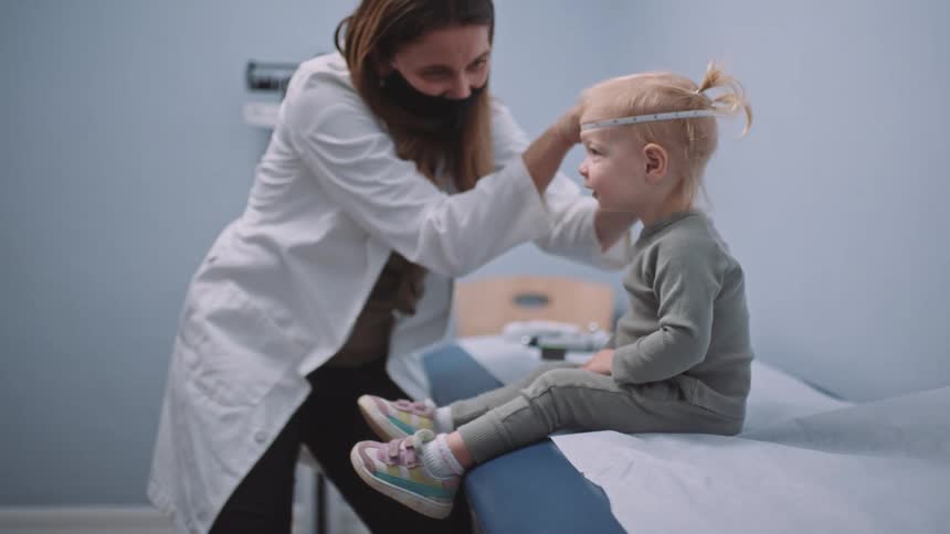 Female doctor wearing a face mask measuring the head of a little girl in her office