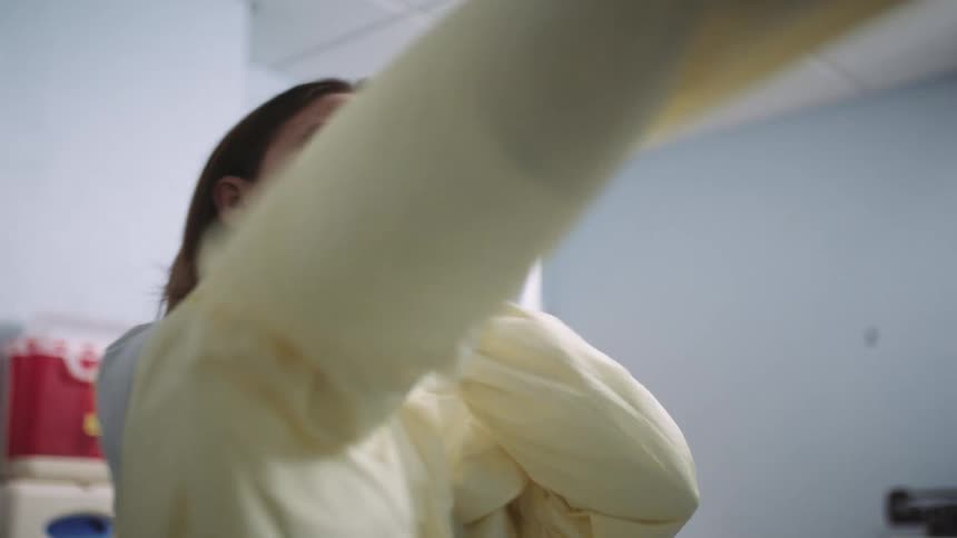 A female medical professional quickly dons a disposable gown to cover he clothing