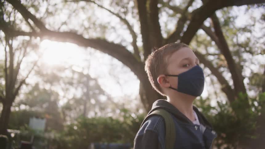 A young boy wearing a face mask walks briskly along a street lined with trees