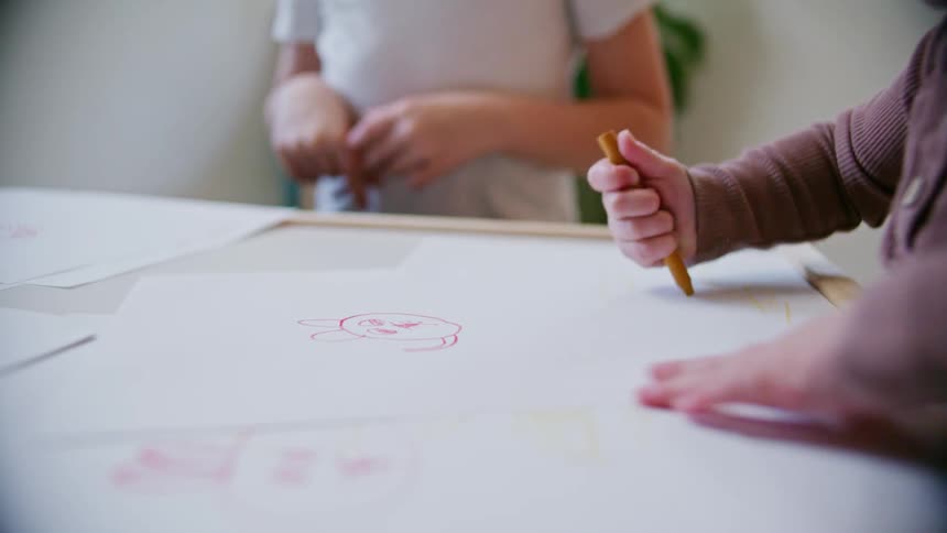 A young holds a crayon and scribbles on paper spread across a table as his brother watches