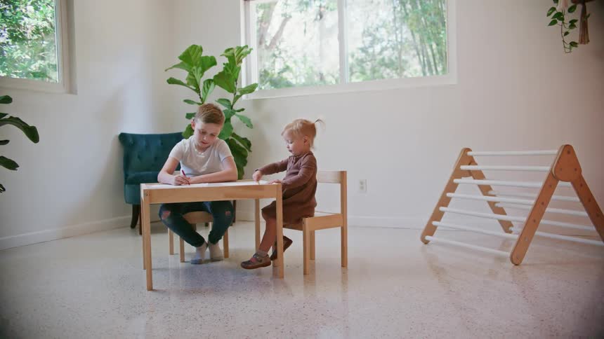 A little boy colors at a small table while his sister laughs