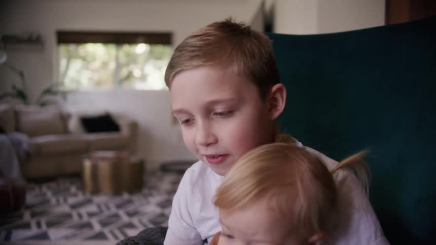 A boy reads a picture book to a small girl sitting on his lap as they sit under a blanket on a chair in the living room of a house during the day