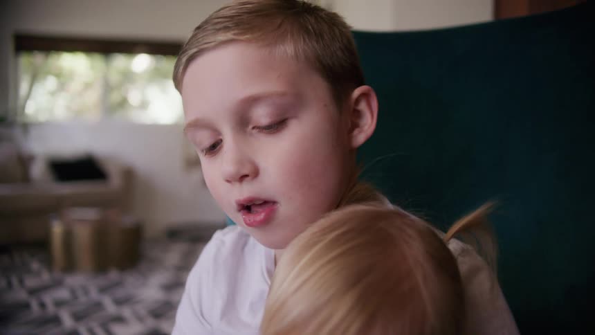 A boy reads a picture book to his younger sister as she sits in his lap in a chair in the living room of a house during the day