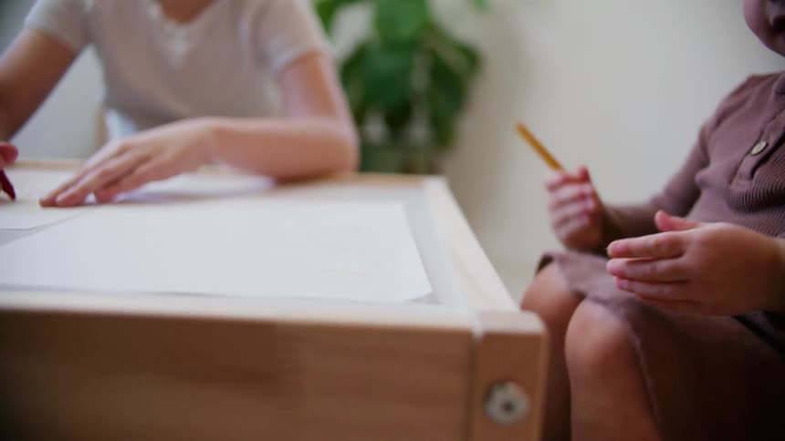 Two children sitting on chairs at a desk writing on paper with colored pencils
