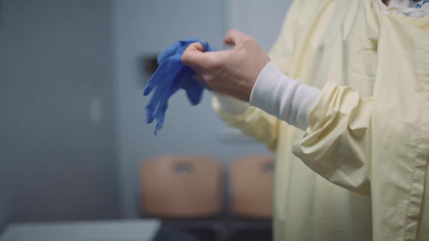 A female medical professional in a disposable gown puts on a pair of disposable gloves