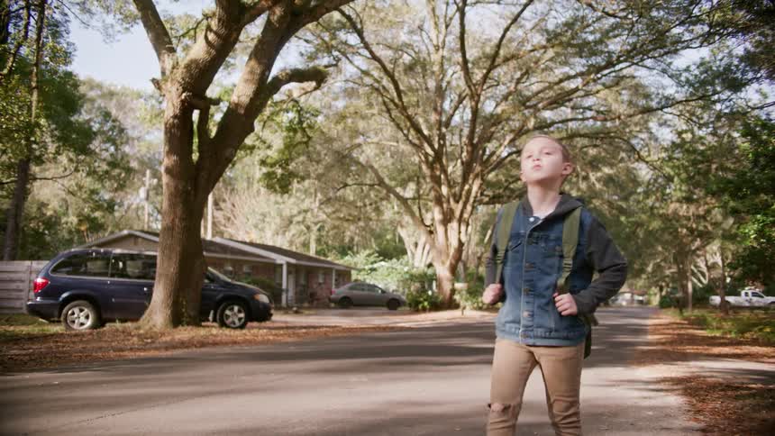 A young boy with a backpack casually walks along street in a residential neighborhood