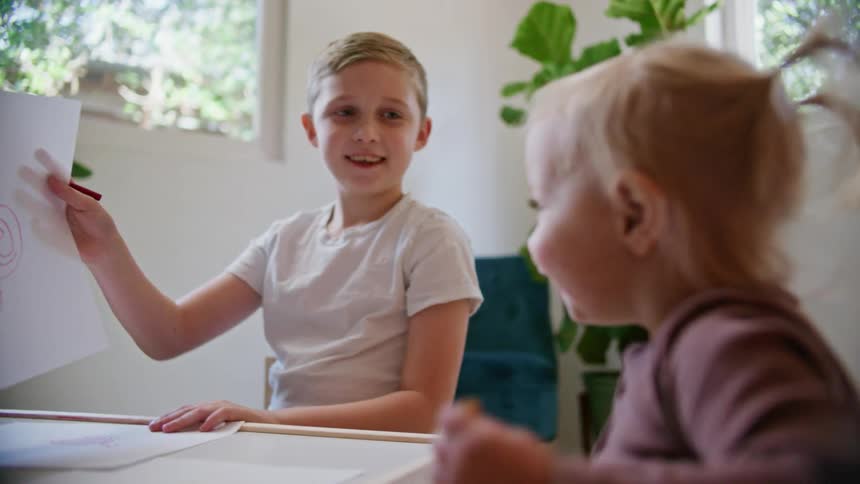 A boy lifts his crayon drawing from a table to show his younger sister