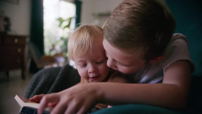 A boy shows a book to a small child sitting in his lap on a chair in the living room of a house during the day