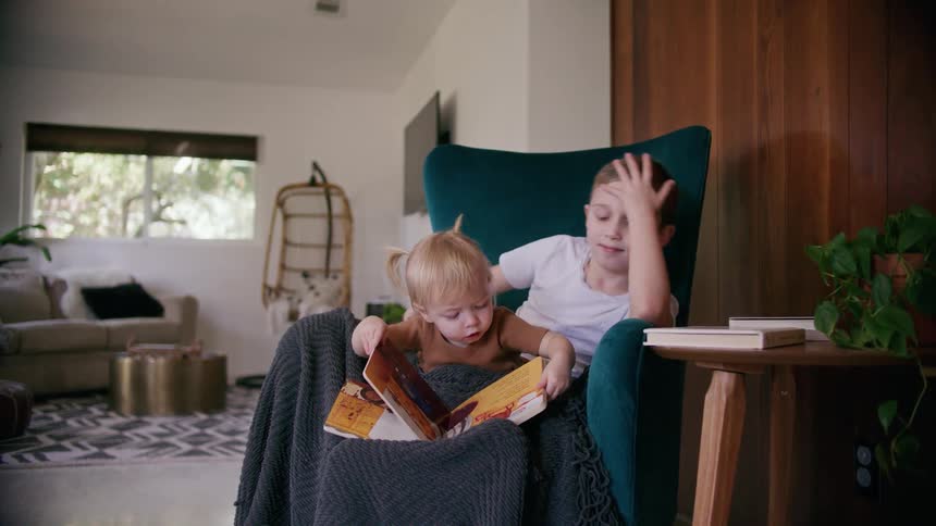 A little girl sits on her brothers lap at home and looks at a book as he plays with her pigtails and points at pictures for her