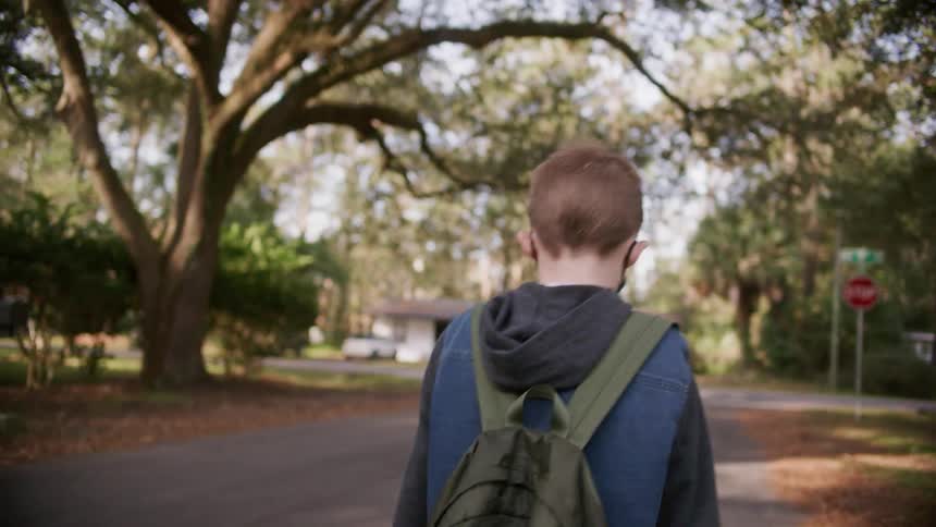 A young boy wearing a backpack walks along a road in a residential neighborhood