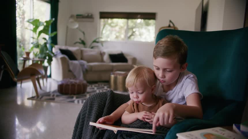 A boy holds his sister in his lap and reads to her as they sit in a large green chair at home