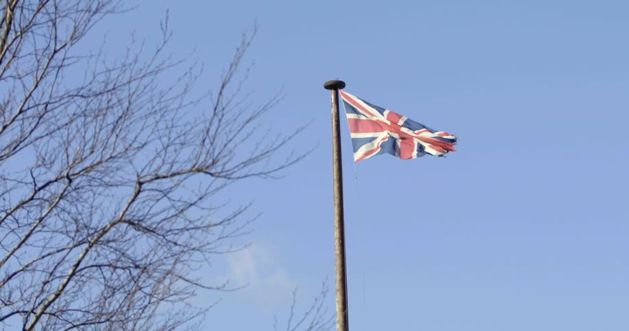 A flag for the united kingdom flies on top of a large flag pole near tall barren trees