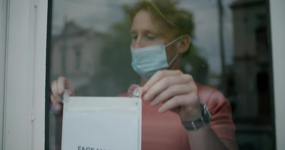 A business owner hangs a sign in the door doorway stating that face masks are required