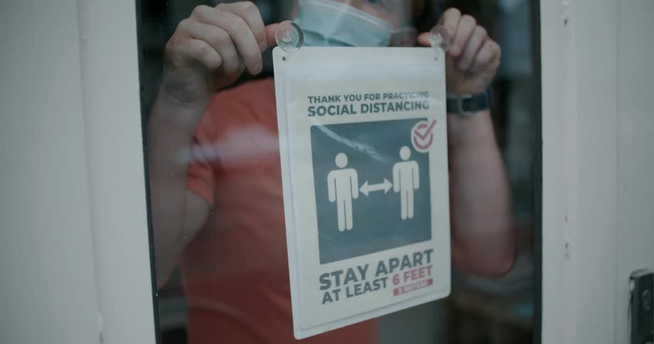 A man in a medical mask posts a social distancing sign in the window of a shop in a city during the day