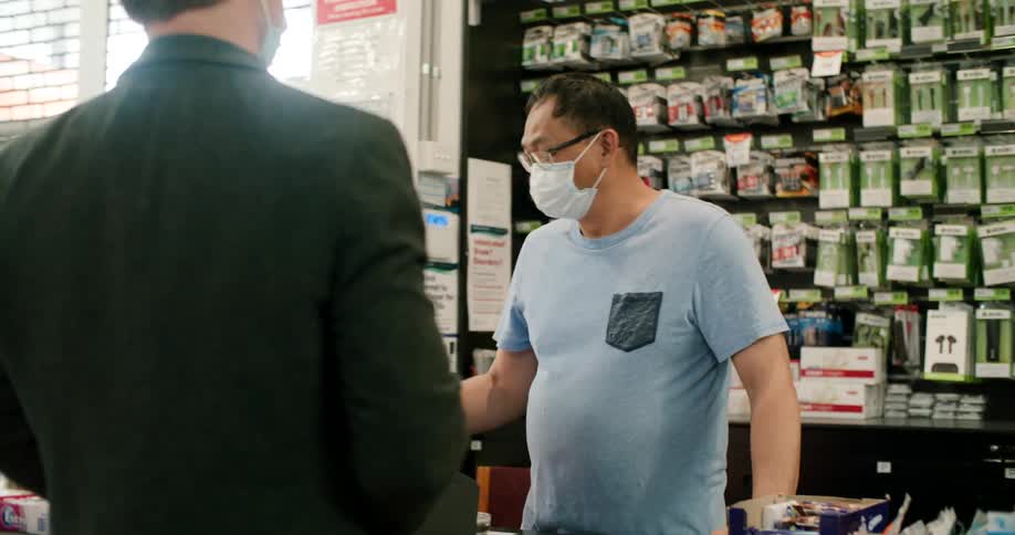 A masked customer hands a pair of bananas to a masked cashier at the checkout of a shop during the day