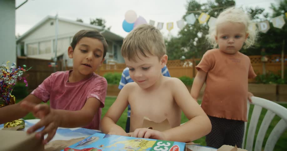 A little girl stands on a chair behind her brother as he opens presents at his birthday party in the backyard