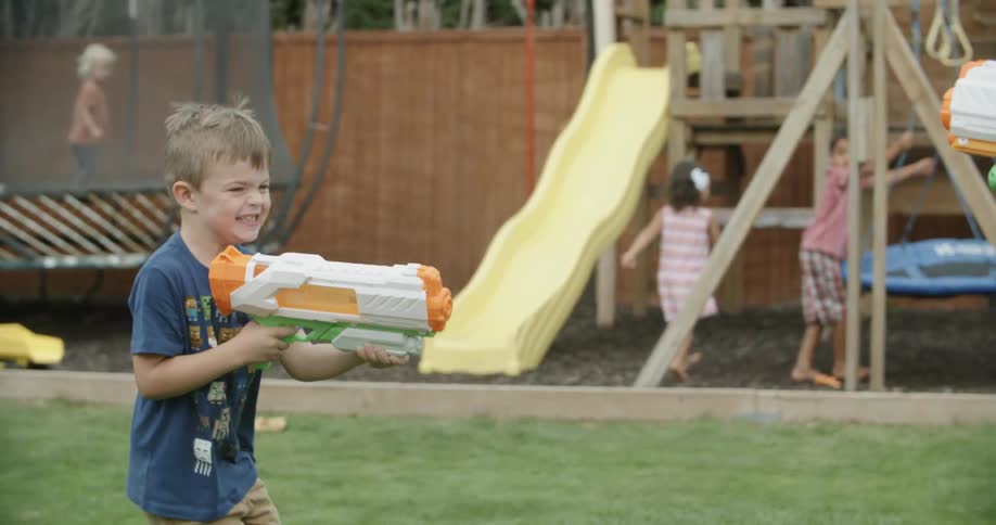 Two boys in a playground playing with water guns with children a trampoline and slides in the background