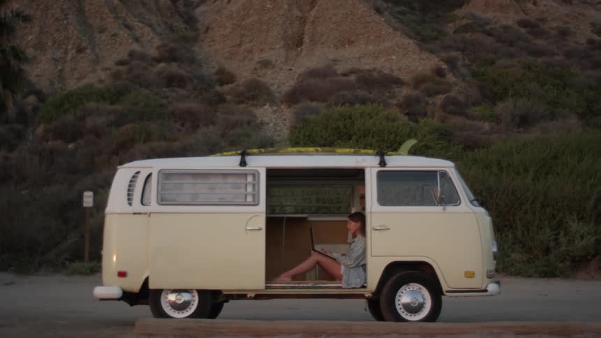 Young woman sitting on the floor of a volkswagen van parked on a rural road working on a laptop