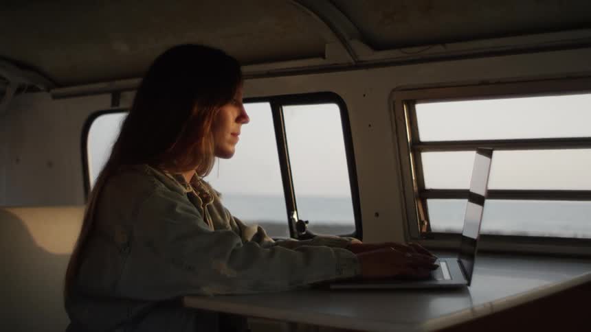 A woman sits in a small van parked next to the ocean working on a laptop
