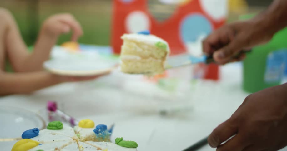 Children sit at a table and hold plates as a person serves a slice of birthday cake
