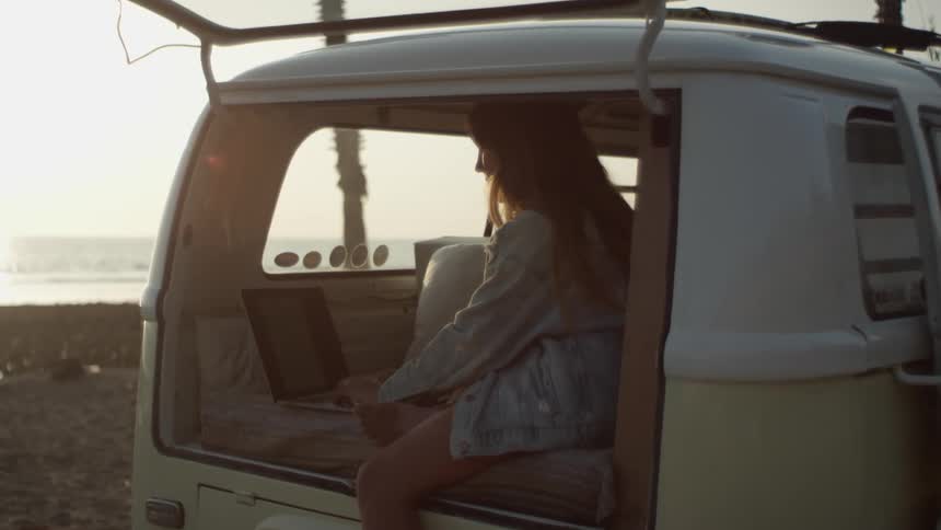 A young woman types on a laptop while sitting in the back of a van parked on a beach with its rear door open