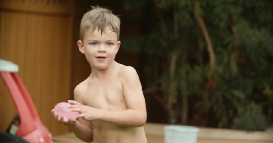 A young shirtless boy playing with water balloons in the backyard at home