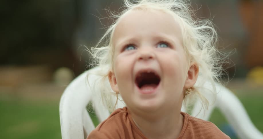 Young blonde girl gesturing happily sitting in a plastic chair over the lawn in the backyard