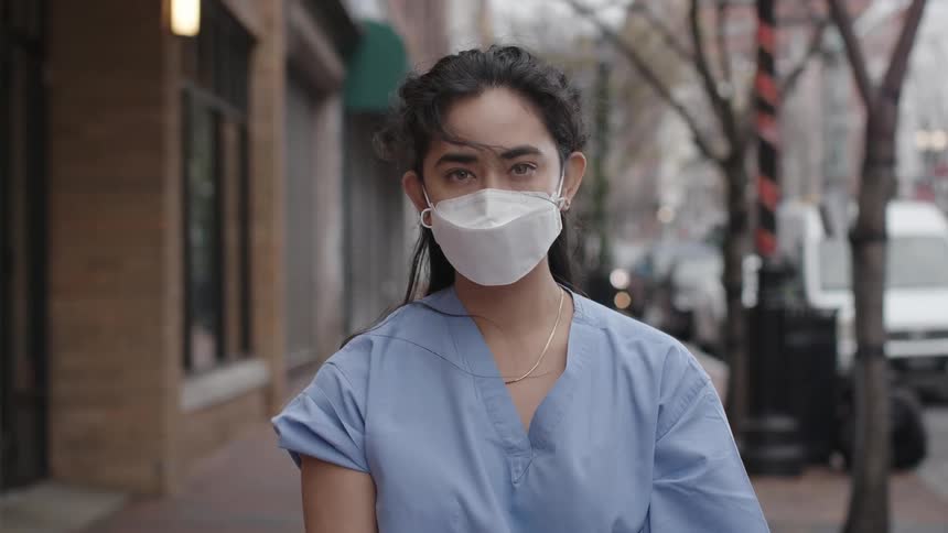 A nurse poses in blue scrubs and a face mask while standing on a city sidewalk