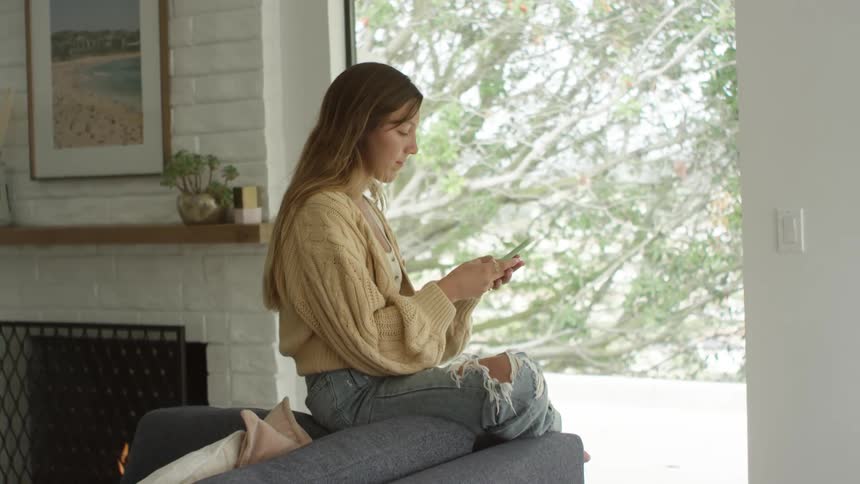 A young woman in ripped jeans sits on the back of a couch and texts