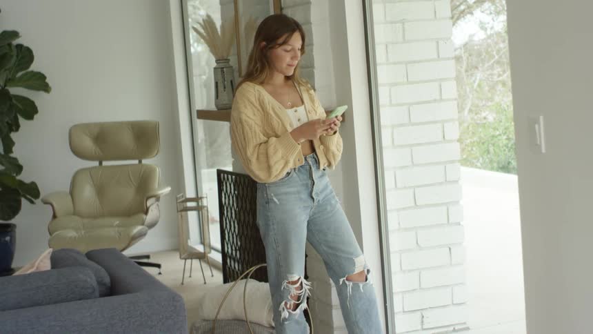 A woman in ripped jeans typing on a cell phone while standing next to a living room window