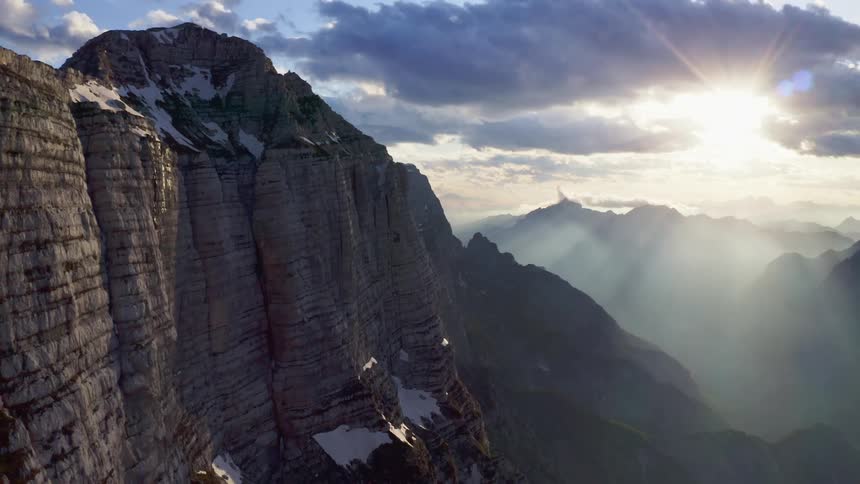 Aerial view of sunlight filtering through clouds shining over the cliffs of a snow covered mountain range