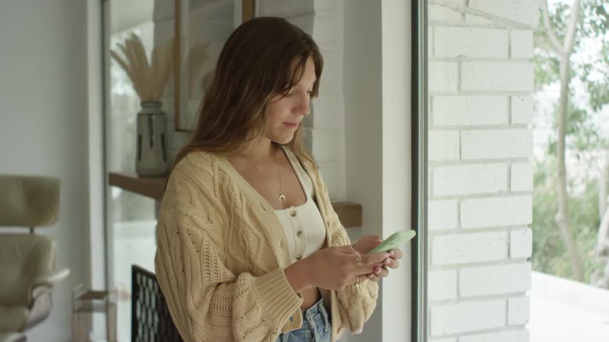 Young woman texting on a smartphone in the living room at home next to a window