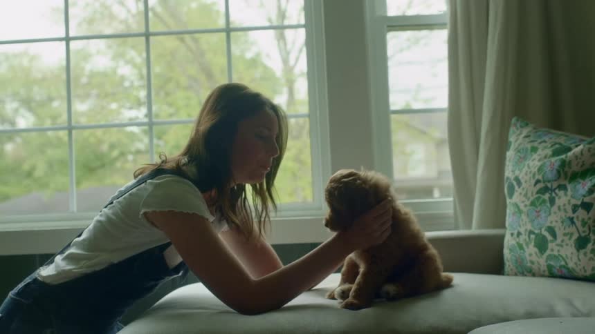 A woman petting a puppy on a bed with a window and curtains in the background