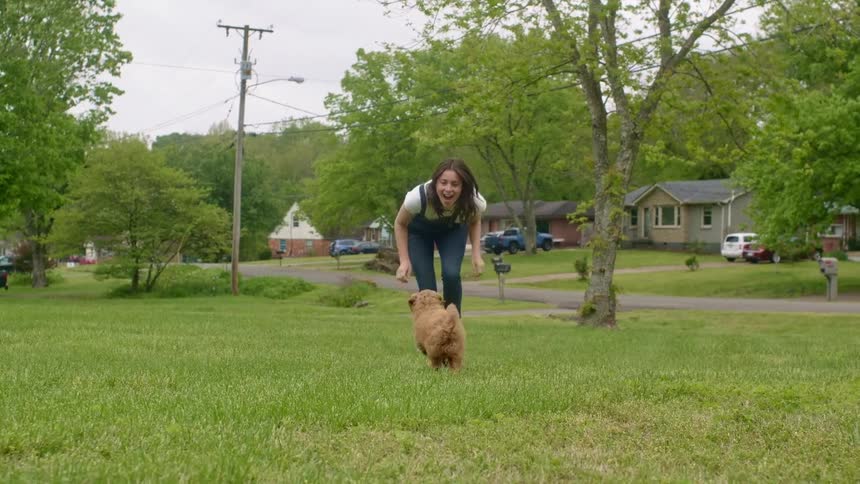 A woman playing with a puppy on grass with houses trees the sky and clouds in the background