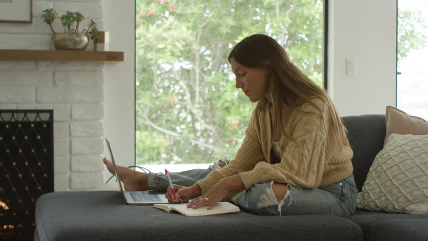 A young woman sitting on a sofa writes in a notebook while looking at the screen of a laptop in the living room of a house during the day