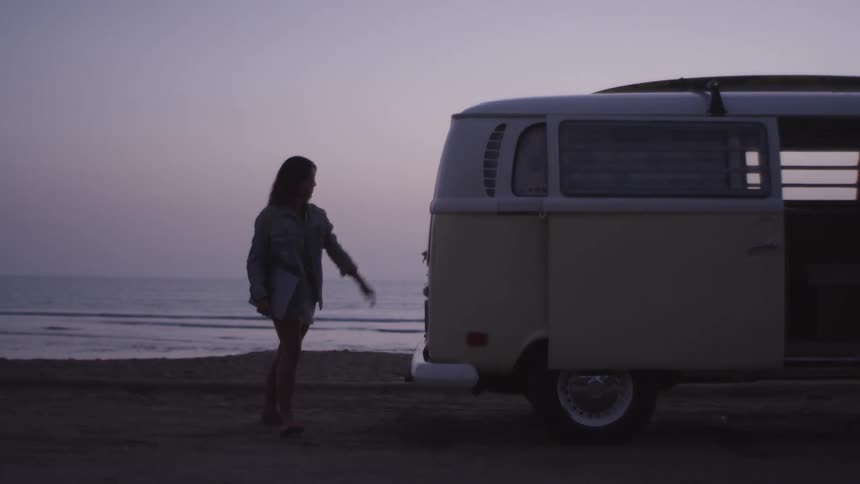 A young woman takes a laptop out of the back of her van that is parked on the beach and closes the back