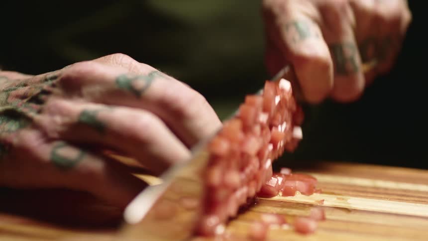 Close up of a tattoed man cutting tomatoes with a sharp kitchen knife over a wooden board