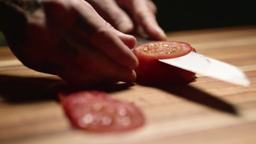 A man uses a sharp knife to slice a tomato on a cutting board