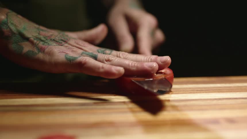 A man with tattoos on his hands and fingers slices a tomato paper thin