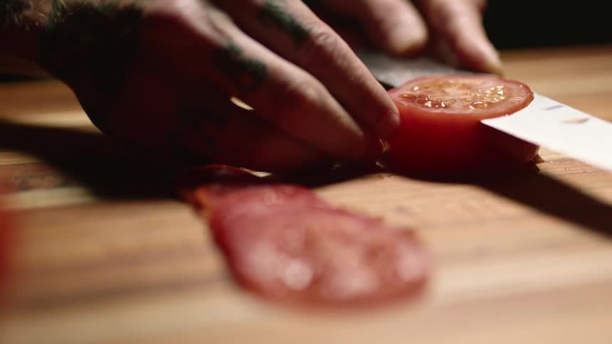 A person uses a knife to cut a tomato into thin slices on a wooden cutting board