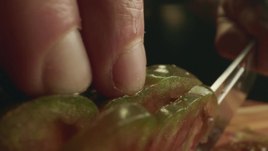 A person carefully holds a tomato while using a knife to cut very thin slices
