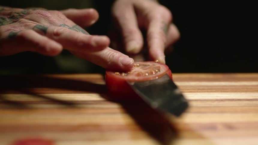 A chef thinly slices a tomato on a wooden counter