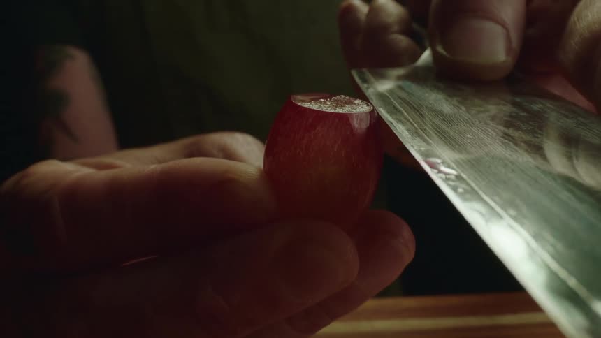 A person cutting a grape with a knife over a cutting board