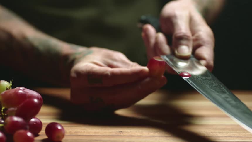 A man holds a grape in his hand and slices it with a large knife