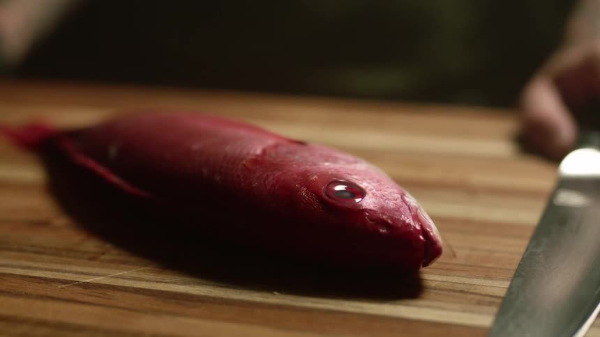 A red fish lies on a wooden cutting board as a person holds a knife in preparation for cooking